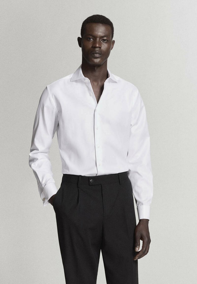 Jeune homme portant une chemise blanche boutonnée au col ouvert et un pantalon noir, debout avec une main dans la poche devant un fond uni.