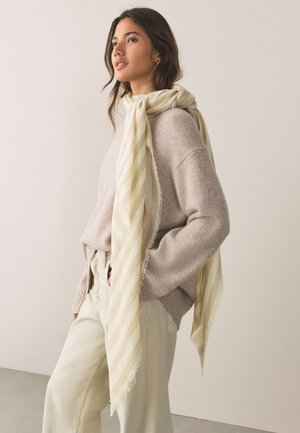 Vrouw draagt een lichtbeige trui en crèmekleurige wijde broek, met een lange, bleekgele gestreepte sjaal losjes om haar nek gedrapeerd.