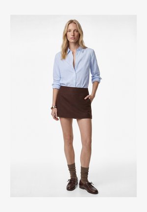 Kvinde iført en blå stribet skjorte med knapper, brun mininederdel, posede brune sokker og brune læder snørestøvler, stående med den ene hånd i lommen.