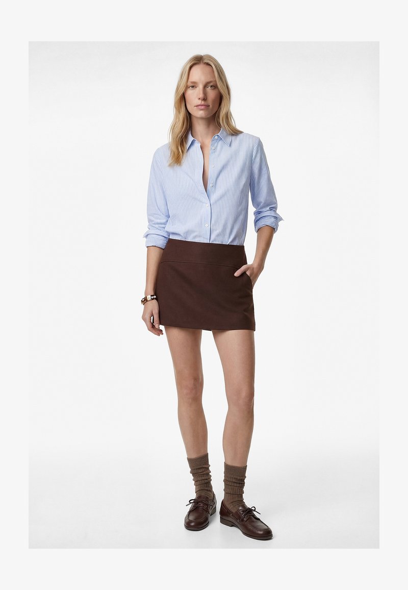 Mujer con camisa de botones de rayas azules, mini falda marrón, calcetines marrones holgados y zapatos marrones de cuero con cordones, de pie con una mano en el bolsillo.