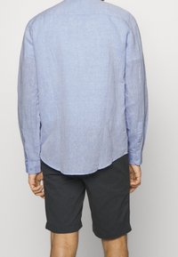 Camicia azzurra chiara a maniche lunghe con design casual, polsini con bottoni e orlo arrotondato, abbinata a pantaloni corti neri. Tessuto misto visibile.