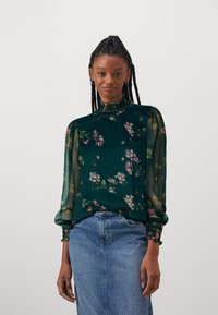 Blusa floreale verde con maniche trasparenti, collo alto e polsini arricciati. Abbinata a una gonna di denim, che presenta una vestibilità comoda e un motivo floreale.