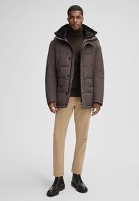 Giacca imbottita marrone con colletto in pelliccia sintetica, con chiusura a bottoni, tasche laterali e taglio dritto, abbinata a pantaloni in corduroy beige e stivali neri.