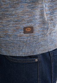 Suéter de punto en tonos azules y marrones con un dobladillo acanalado. Presenta una etiqueta de cuero marrón con el logo "Petrol Industries". Combinado con unos vaqueros de denim oscuros.