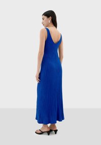 Robe longue bleu à côtes avec un dos en V, présentant un haut cintré et un bas évasé. Associée à des sandales à talons ouverts noires.