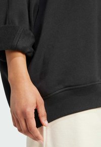 Svart sweatshirt med ribbad kant och muddar, som har en avslappnad passform. Synlig struktur och mjukt tyg, kombinerat med ljusa byxor.