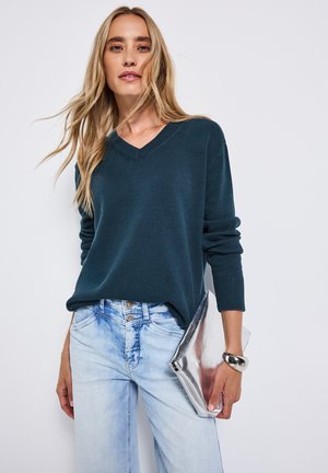 Pull v-neck en bleu canard avec des poignets côtelés assorti à un jean bleu clair. Sac à main en métal argenté et coupe large en denim.