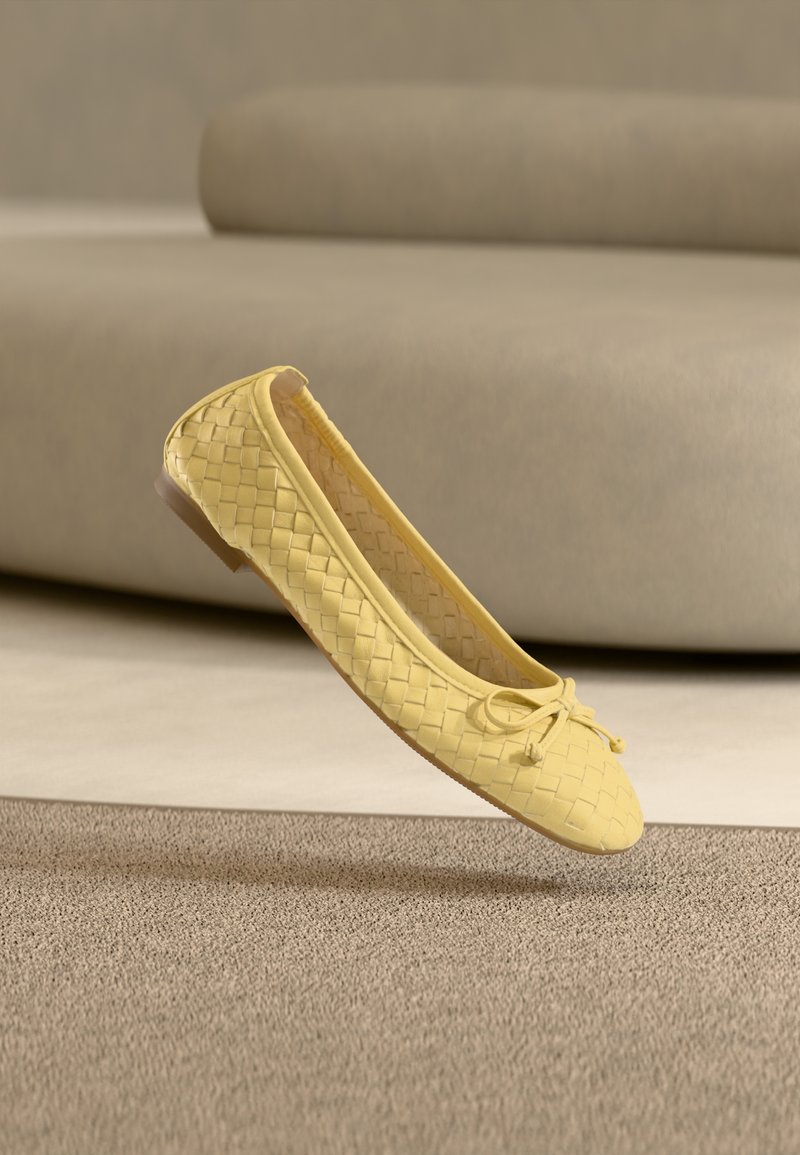 Ballerine jaune tissée avec un petit nœud, flottant au-dessus d'un tapis beige devant un canapé de couleur claire.