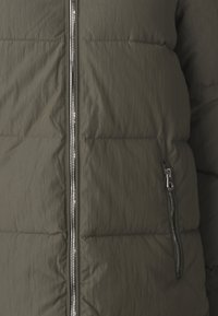 Veste en duvet vert olive avec un motif de piquage horizontal, fermeture éclair à l’avant et détails de poches zippées sur les côtés. Le tissu texturé offre une apparence légère.