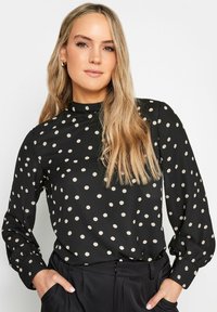 Svart blus med lång ärm, hög halslinning och beige prickar. Tyget verkar slätt med en lätt lyster. Styler med svarta byxor.