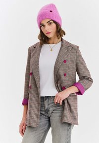 Hnedé kockované sako s ružovými gombíkmi a manžetami, v kombinácii s bielou košeľou a sivými džínsami. Na hlave jasne ružová pletená čiapka.