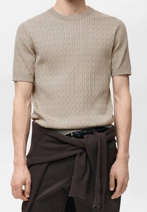 Mand iført beige kortærmet striktrøje, mørke bukser og mørk sweater bundet om livet, kun arme og overkrop vises.