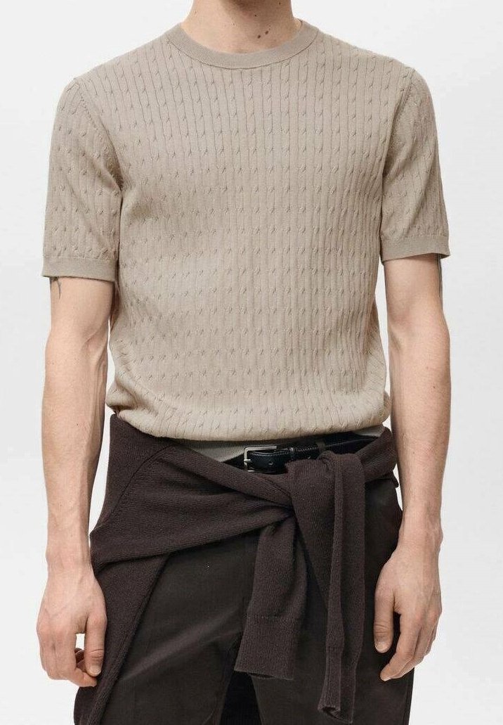 Homme portant un pull en maille beige à manches courtes, un pantalon foncé et un pull foncé noué autour de la taille, seuls les bras et le torse sont visibles.