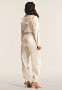 Survêtement en satin ivoire avec une veste à capuche et une taille élastique ; les pantalons se resserrent aux chevilles. Porté avec des sandales à talons transparents.