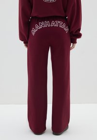 Pantalones de chándal burdeos con un ajuste relajado, con bordado en blanco del texto "MANHATTAN" en la cintura. Fabricados en un tejido suave con una textura lisa.