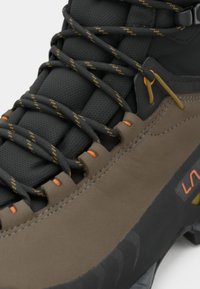 Botas de caminhada castanhas e pretas com língua acolchoada, atacadores com padrão preto e amarelo, e sola robusta projetada para uso ao ar livre.