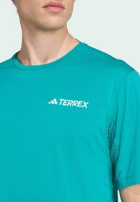 Turkos idrottströja med rund halsringning, tillverkad av lättviktsmaterial. Har en vit "TERREX"-logotyp på framsidan och texturerade detaljer på sidorna.