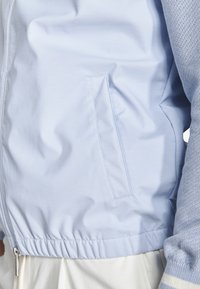 Hellblaue Jacke mit Seitentasche und elastischem Saum, getragen über einem hellblauen Strickärmel und weißen Hosen.