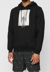 Felpa nera con cappuccio con grafica frontale di una mano che fa il segno di pace, con la parola "pace" sotto. Vestibilità classica con tasca a marsupio.