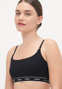 Bralette nera in cotone con spalline sottili regolabili e fascia elastica con il logo "HUGO" bianco. Texture liscia e design minimalista.