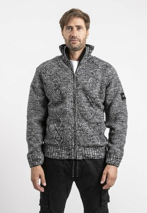 Homme portant un pull en maille gris texturé à fermeture éclair avec un col haut, debout devant un fond blanc uni.