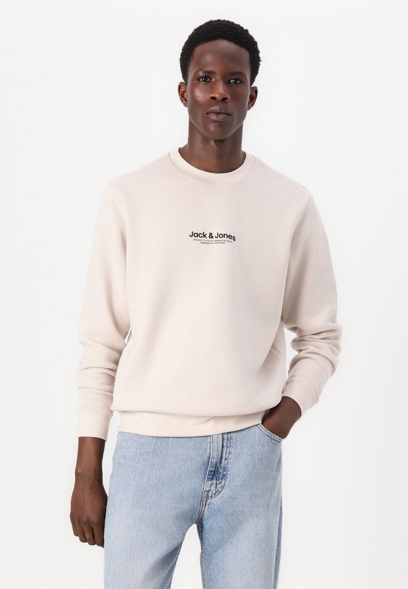 Lichtbeige sweatshirt met ronde hals en lange mouwen. De voorkant toont het zwarte "Jack & Jones" logo. Gecombineerd met lichtblauwe denim jeans.