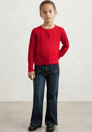 Rote Strickjacke mit einem strukturierten Muster, Knopfverschluss vorne und gerippten Bündchen, kombiniert mit dunkelblauen Schlagjeans und schwarzen Schuhen.
