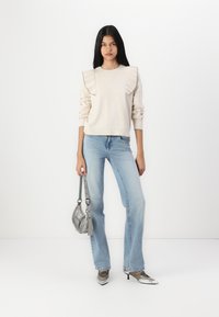 Suéter beige con detalles de volantes plisados en los hombros, combinado con jeans acampanados de color azul claro y zapatos plateados, sosteniendo un pequeño bolso gris.