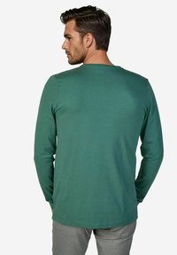 Langärmliges T-Shirt mit Rundhalsausschnitt aus einfarbigem dunkelgrünem Stoff. Glatte Textur und lockere Passform, kombiniert mit grauer Hose. Minimalistisches Design.