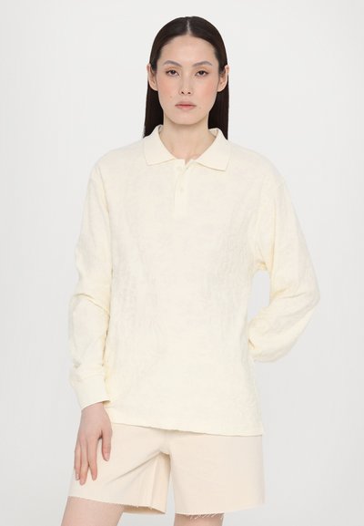 Polo a maniche lunghe in morbido tessuto crema, con colletto classico e una texture a rilievo. Indossato con pantaloni corti beige chiaro.