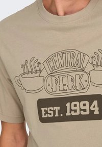 Camiseta beige con cuello redondo que muestra un gráfico marrón de una taza de café y el texto "CENTRAL PERK" y "EST. 1994". Diseño de línea simple.