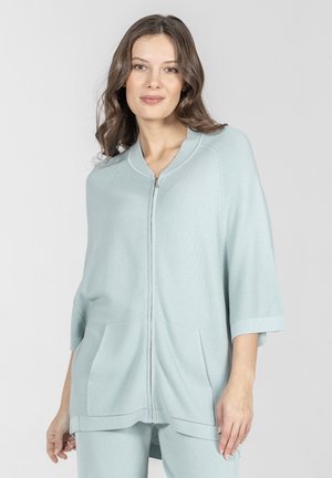 Kobieta z falującymi włosami, ubrana w jasnoniebieski sweter na suwak z rękawami 3/4 oraz pasujące spodnie, stojąca na jednolitym jasnym tle.