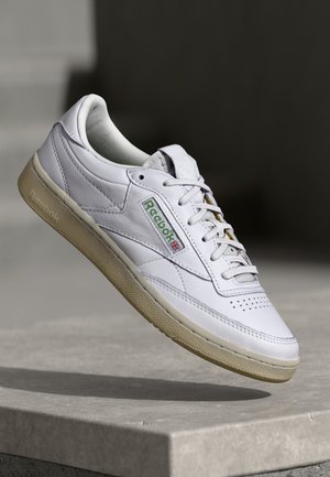 Valkoinen nahkainen lenkkari, jossa on rei'itetty kärki, vihreä ja punainen logo sekä beige kumipohja. Varustettu pyöreillä nauhoilla ja sileällä pinnalla.