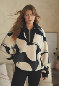 Fliisist valmistatud pullover, millel on kreemjas ja mustas abstraktne muster, tõmblukuga krae ja sooniktehnika aktsendid. Pehme tekstuur ja mugav lõige.