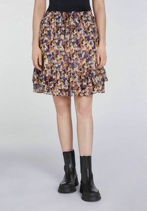 Femme portant une mini-jupe à motif floral avec des volants, associée à des bottines noires épaisses, debout devant un fond uni.