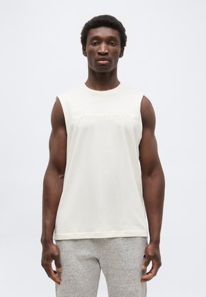 Muž stojaci čelom dopredu, oblečený v bielom tielku značky Champion bez rukávov a svetlosivých teplákoch, na jednoduchom pozadí.