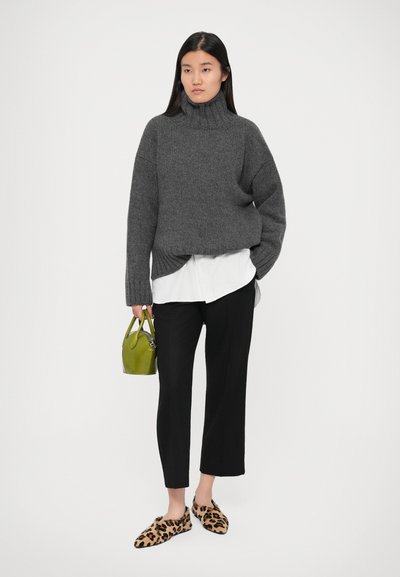 Donna che indossa un maglione grigio a collo alto sopra una camicia bianca e pantaloni neri, tiene in mano una piccola borsa verde con delle ciabatte con stampa leopardata.