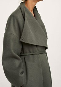 Vrouw in een oversized olijfgroene wollen jas met brede revers en een dunne riem om de taille, staand tegen een effen achtergrond.