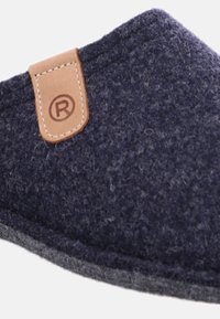 Närbild av en marinblå texturerad toffel med grå sula och en tan läderetikett med registrerat varumärkessymbol.