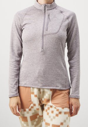 Personne portant un pull à col montant à fermeture éclair trois-quarts manches longues de couleur violet clair et un pantalon en polaire beige à motif avec des patches bruns sur les cuisses.