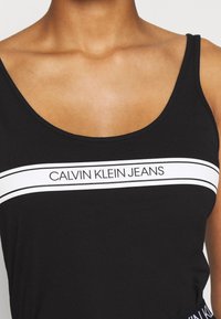 Černý top bez rukávů s širokým výstřihem a bílým logem "CALVIN KLEIN JEANS" přes hrudník, vyrobený z měkké, elastické tkaniny.