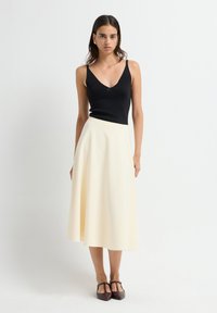 Zwart mouwloos topje met een V-hals, gecombineerd met een vloeiende, knie-lange romige rok. Het model draagt donkerbruine, puntige schoenen.
