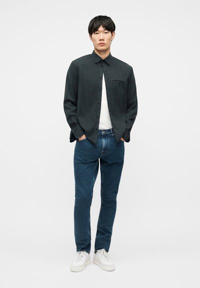 Młody mężczyzna stojący, ubrany w ciemnozieloną koszulę na guziki na białej koszulce, niebieskie dżinsy i białe trampki, na tle gładkiego jasnego tła.