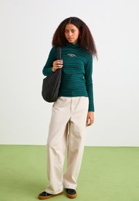 Grøn og marineblå stribet turtleneck top, beige brede bukser, sort quiltet tote taske, sorte sko med brune såler. Krøllet hår.