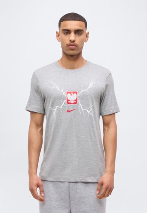 Ung mand iført grå t-shirt med hvid lyn-design, rød Nike-logo og hvid ørnemblem på brystet, stående mod en ensfarvet baggrund.