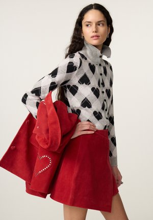 Vrouw draagt een grijze trui met zwarte harten, houdt een rood jasje met hartdetail en een rode rok tegen een effen achtergrond.