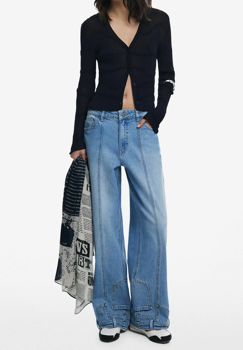 Femme portant un cardigan cropped noir à côtes, un jean bleu à jambes larges avec des détails en patch, des chaussures blanches et noires, tenant un tissu noir et blanc à motifs.