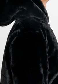 Sort faux fur hoodie med en blød, plysset tekstur, der har en hætte og lynlås. Stofmet har en glat, blank overflade og en afslappet pasform.