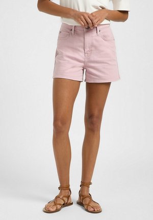 Vrouw draagt lichtroze denim korte broek, wit topje, en bruine sandalen met bandjes, staand met handen gevouwen voor zich.