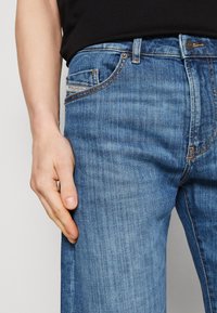 Vaqueros de mezclilla azul con un corte slim, que cuentan con un bolsillo para monedas, costuras naranjas y un cierre de botón metálico. Textura suave con ligero desvanecimiento.